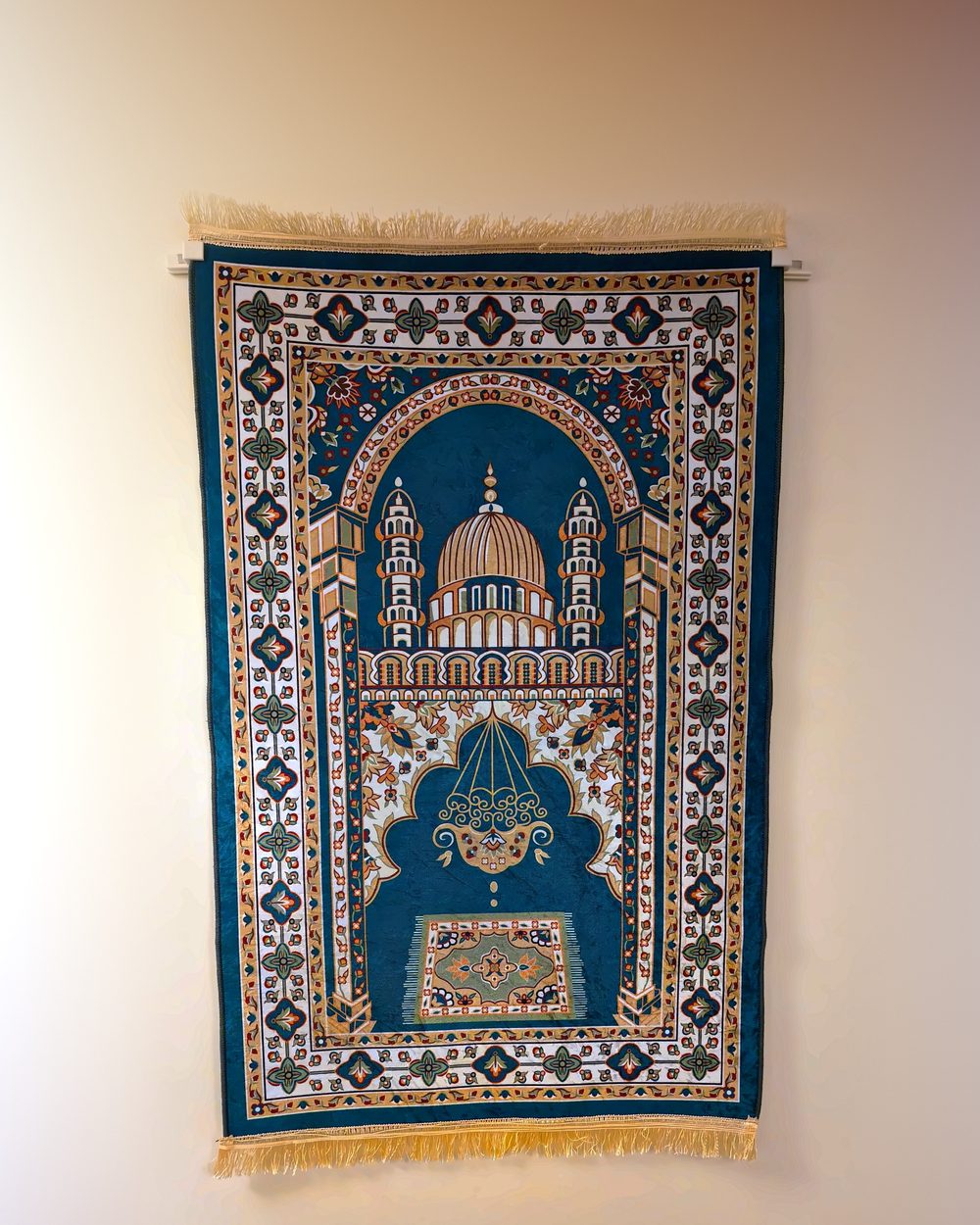 A teal and gold prayer mat hanging flat on a cream wall, held at its top corners by two small ivory MatSnap clamps